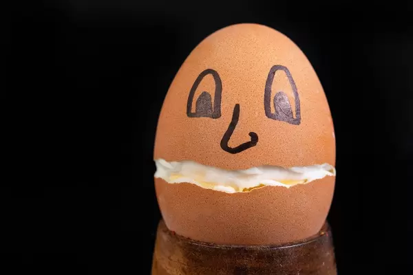 Hartgekochtes, in der Mitte gebrochenes Ei mit aufgemaltem Gesicht vor schwarzem Hintergrund