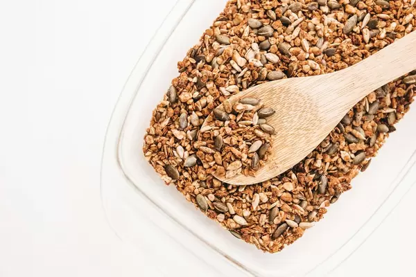 Hausgemachtes Granola aus Kernen, Samen und Nüssen mit Holzlöffel in weißer Schüssel auf weißem Hintergrund