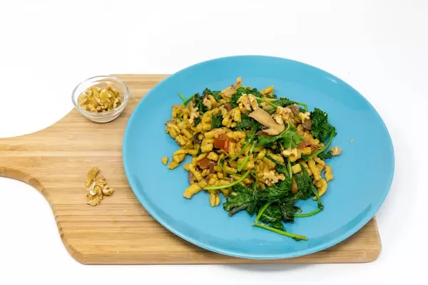 Hello Fresh - Herbstliche Grünkohl-Spätzle-Pfanne in cremiger Soße mit gerösteten Walnüssen auf einem blauen Teller auf einem Holzbrett auf weißem Hintergrund