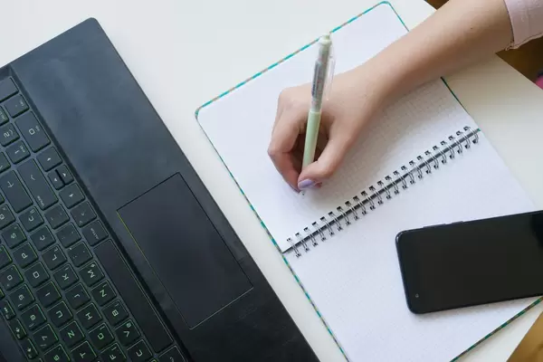 High angle shot of a womans hand taking notes on a notebook, with laptop and smartphone aside