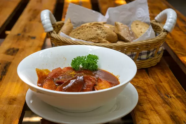 Homemade Currywurst with Spicy Tomato Sauce, Curry Powder and Parsley on a Wooden Table with a Basket with Bread Slices in a German Restaurant