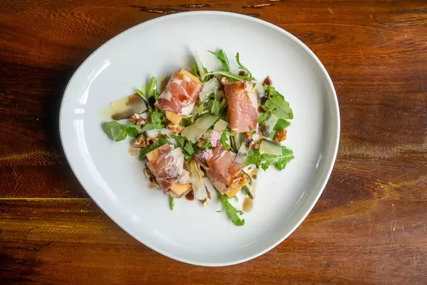 Honigmelone in Parmaschinken mit Parmesan, Rucola und Balsamico auf einem weißen Teller von oben auf einem Holztisch fotografiert