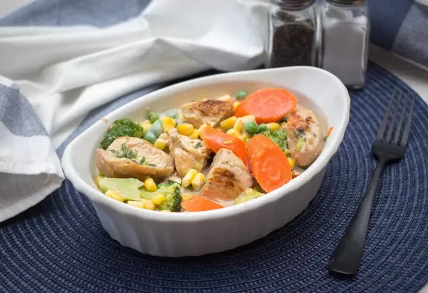 Hühnchen Kasserolle mit Karotten, Mais, Brokkoli und Erbsen in weißer Porzellanschale auf blauem Tischset mit Salz und Pfeffer im Hintergrund