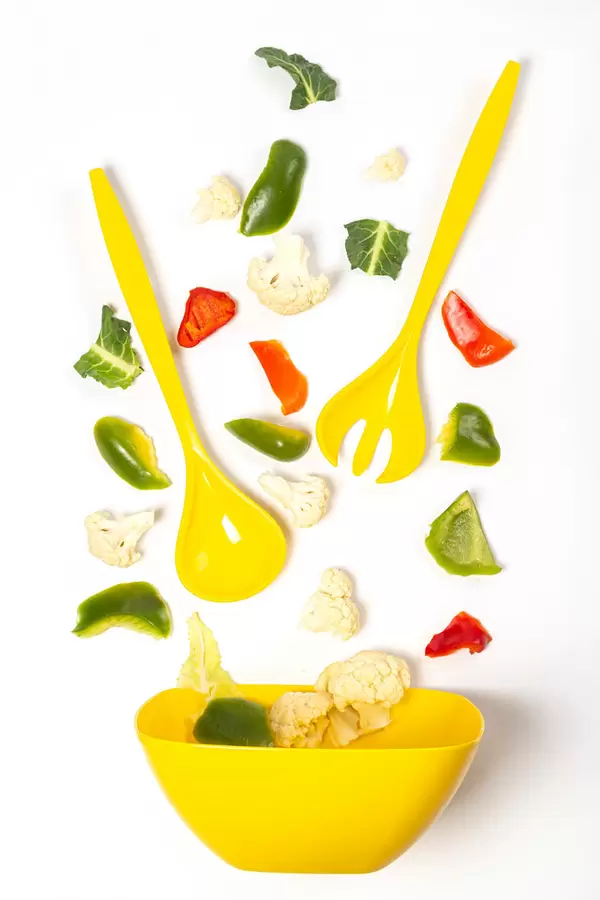 Ingredients for vegetable salad scattered on white background with yellow salad bowl, spoon and fork