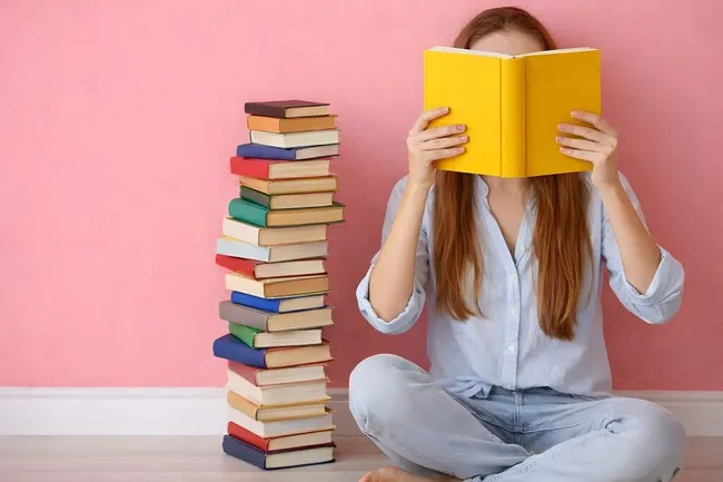 Junge Frau liest Buch neben Bücherstapel vor rosa Wand