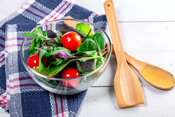 Kalorienarmer Salat mit frischen Tomaten und Kräutern in einer Glasschale auf blauem Küchentuch neben Holzbesteck
