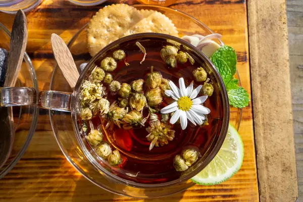 Kamillentee in einer Glastasse auf einem Glasuntersetzer mit Holzlöffel, Keksen, Zitronengras, Minze und Zitrone von oben auf einem Holzbrett fotografiert