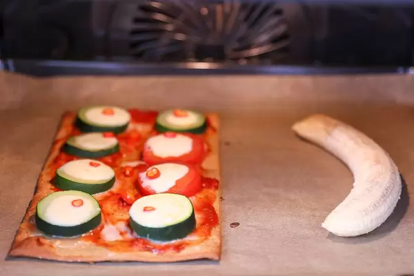 Käse überbackene Zucchini-Scheiben mit Tomatensoße auf Pizzateig neben geschälter Banane im Backofen - Nahaufnahme frontal