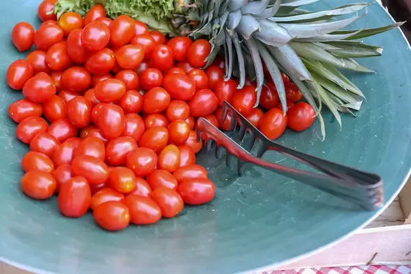Kleine, reife Tomaten liegen mit einer Salatgabel und neben einer Ananas auf einem türkisen Teller