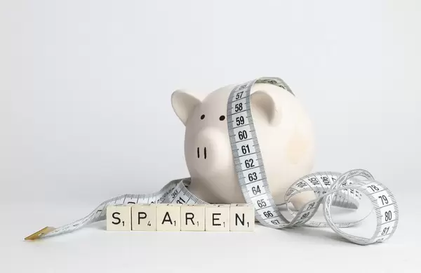 Kleine Schweine-Sparbüchse umgeben von Bandmaß und hinter Scrabble-Würfel welche 'Sparen' buchstabieren - frontale Nahaufnahme