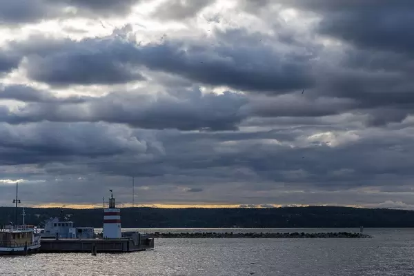 Kleiner weiß-roter Leuchtturm bei bewölkter Abenddämmerung im Hafen von Lahti, neben einem Hausboot, am Vesijärvi-See