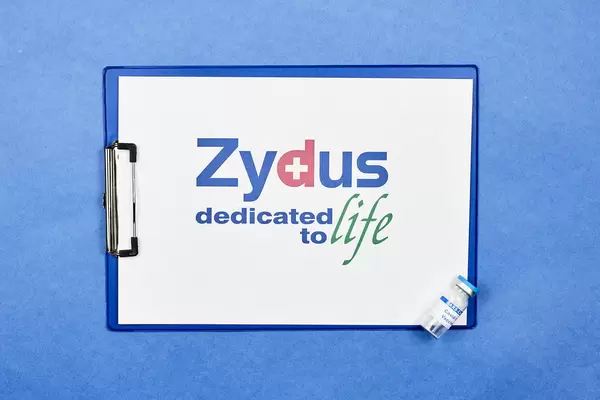 Klemmbrett vor blauem Hintergrund mit Zydus Pharmaceuticals Logo und Corona-Impfstoff Ampulle