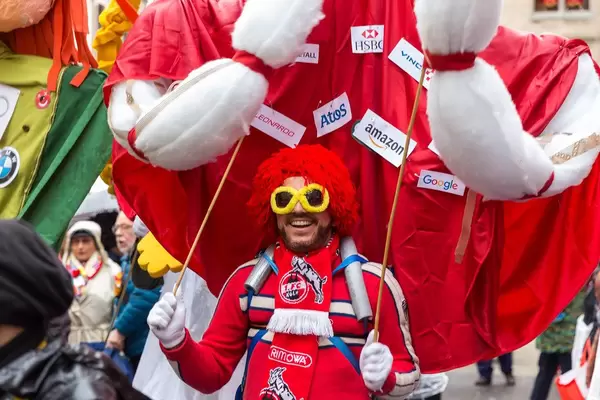 Kölner Jeck mit Perücke beim Zoch vor dem Zoch trägt die Figur "Seine Monströsität Prinz Narren-Kapitalismus" auf dem Rücken