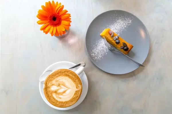 Kürbiskuchen mit Kernen und Puderzucker verziert auf einem grauen Teller mit einer Tasse Cappuccino und einer orangenen Gerbera