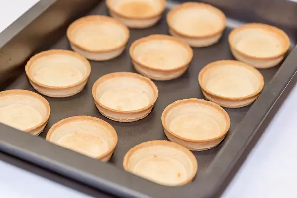 Leere Törtchenschalen aus Mürbeteil auf Backblech vor weißem Hintergrund