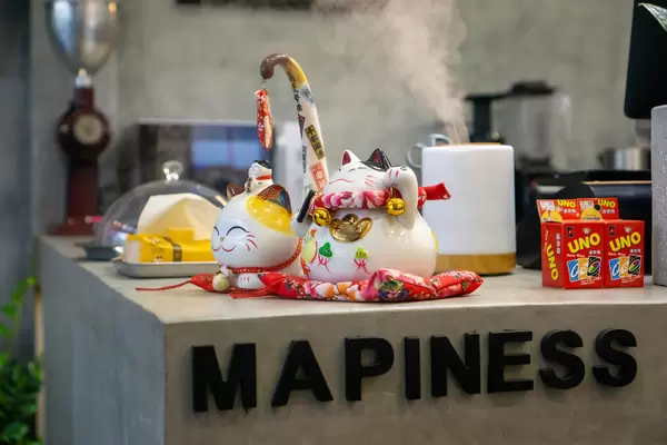Lucky Cats, Moisturizer and Uno Playing Cards on a Counter with Cafe Equipment in the Background