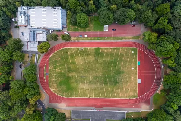 Luftaufnahme eines Sportplatz mit Laufbahn und Wiese, neben der Trainingshalle des ASV Köln, umgeben von Wald