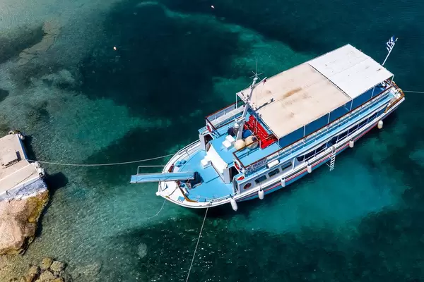 Luftbild einer blau-weißen Fähre mit Steg, im grünen Wasser des Argolischen Golfs, vor der Felsenküste von Spetses, Griechenland