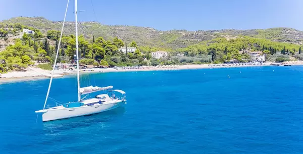 Luftbild eines Segelbootes auf dem klaren blauen Myrtonischen Meer, mit dem Agii Anargiri  Strand von Spetses im Hintergrund