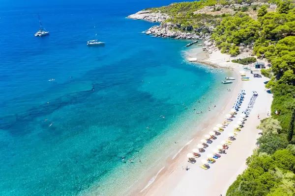 Luftbild von Touristen auf aufgereihten, gelben Sonnenliegen unter Sonnenschirmen, am Agii Anargiri Strand auf Spetses, am blauen Meer