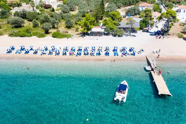 Luftbild zeigt ordentlich aufgestellte Sonnenschirme in blau und weiß, am Sandstrand von Agii Anargiri  und ein Motorsportboot auf dem klaren grünen Meer