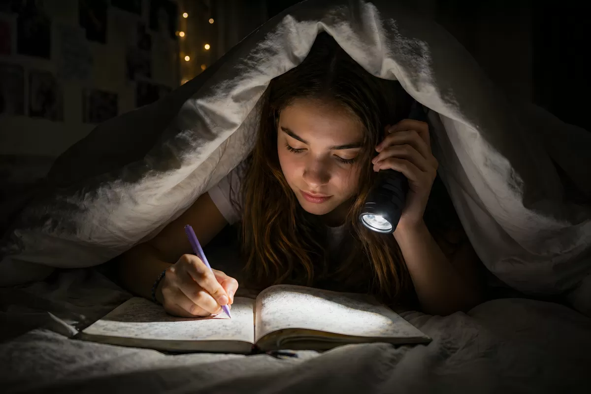 Mädchen schreibt nachts mit Taschenlampe unter Decke