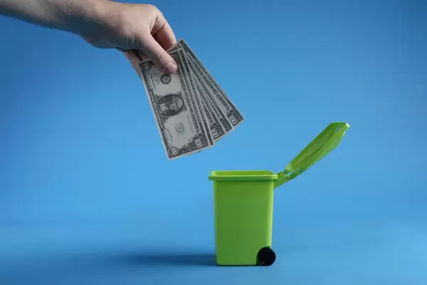 Male hand throwing bundle of money dollars into green trash bin on blue background