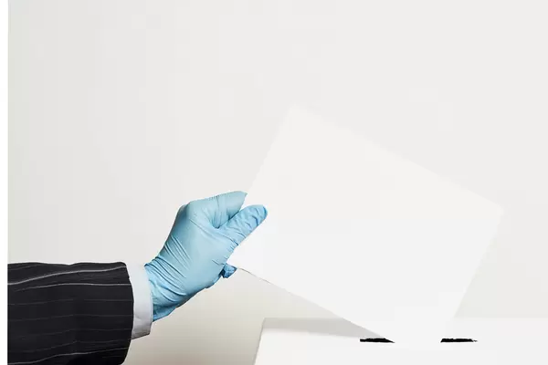 Male hand wearing blue protective latex glove placing ballot paper in vote box