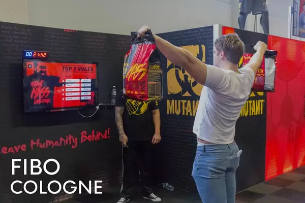 Man at a sports fair does strength training with packages of Muscle Mass Gainer, next to picture title "Fibo Cologne"