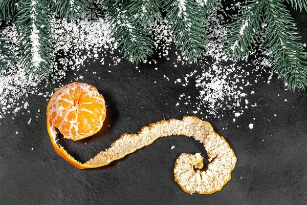 Mandarine mit kunstvoller geschälter Schalte vor verschneitem Tannenbaum auf schwarzer Oberfläche