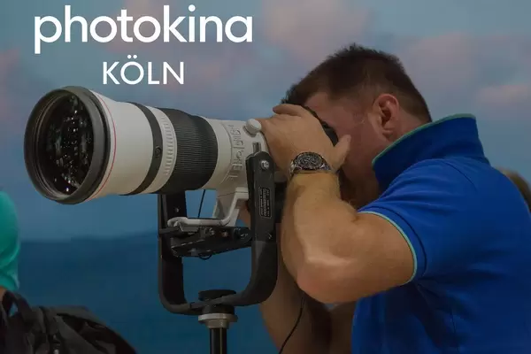 Mann blickt durch ein professionelles und gigantisches Objektiv auf einem Manfrotto-Stativ, neben dem Bildtitel "Photokina Köln"