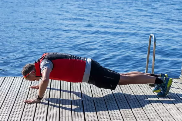 Mann macht Liegestütze während des morgendlichen Work-Outs, auf einem Holzsteg mit blauem Wasser im Hintergrund