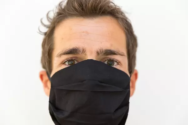 Mann mit schwarzem Mehrweg-Mundschutz, der nur die Augen sichtbar lässt: Nahaufnahme Porträt vor weißem Hintergrund