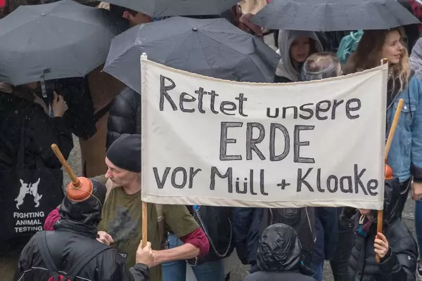 Mann mit Toilettenbürste am Kopf hält einen Schild 'Rettet unsere ERDE vor Müll + Kloake'