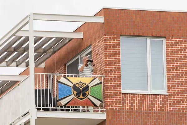 Mann schaut mit Fernglas aus einem Balkon, auf dem eine Flagge mit dem Logo von Tomorrowland Festival aufgehängt ist