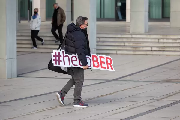 Mann trägt ein Schild mit Schrift #helloBER: Hashtag zur Eröffnung des neuen Flughafens BER in Berlin