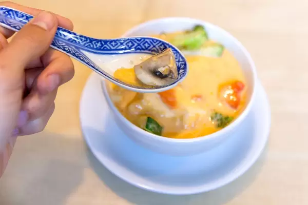 Männerhand löffelt asiatische Kokosmilchsuppe mit Tofu, scharfen Gewürzen und gesundem Gemüse als fleischlose Mahlzeit, in einer Suppenschale