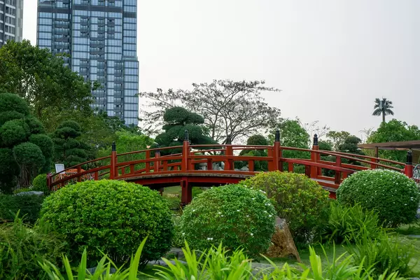 Many Plants and Flowers around a Red Wooden Bridge over a Fish Pond with Vincom Landmark 81 Skyscraper in the Background at Vinhomes Central Park in Saigon, Vietnam