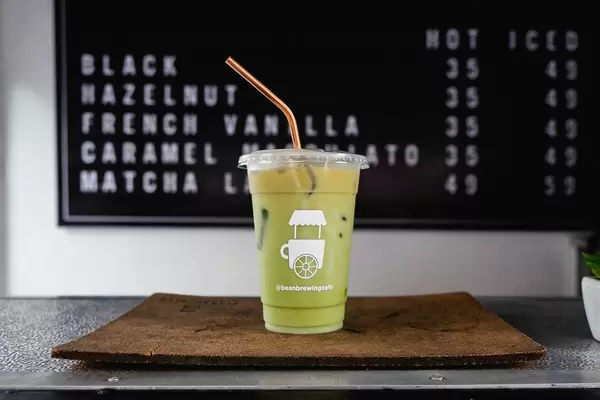 Matcha Ice Latte auf einem brauen Tablett mit dem Kaffeeangebot des Beanbrewing Cafés im Hintergrund