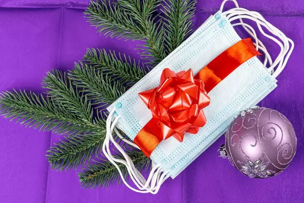 Medical protective face masks with red gift ribbon, Christmas ball and fir branches on a purple background