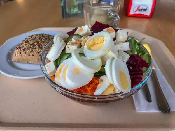 Menu in der Kantine bestehend aus gemischtem Salat mit Karotten, Gurken, Rucola, roter Bete, Fetakäse und Ei, dazu ein Vollkornbrötchen und Tee, bereitgestellt mit Besteck auf beigem Tablett