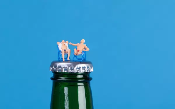Miniature people relaxing in deck chairs on beer bottle on blue background