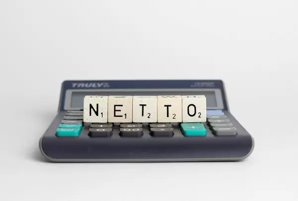 Mit Buchstabenwürfeln gelegtes Wort 'Netto' auf einfachen Taschenrechner in Frontalansicht auf weißem Hintergrund