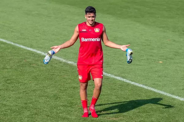 Nadiem Amiri in red Barmenia football jersey, posing and walking barefoot with his blue football shoes in his hands