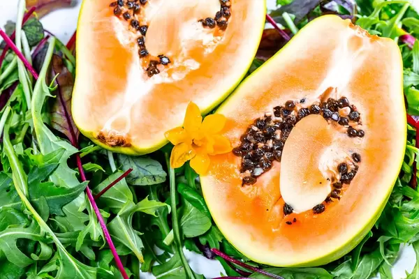 Nahaufnahme einer aufgeschnittenen Papaya Frucht mit Kernen, auf grünem Salat, mit einer Narzisse dekoriert