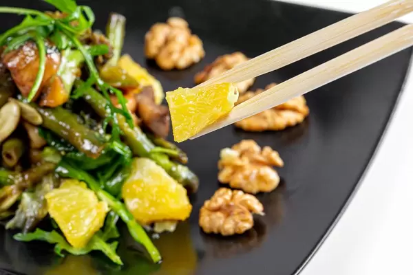 Nahaufnahme eines Stück Orange auf Essstäbchen, vor einem vegetarischen Salat mit Nüssen