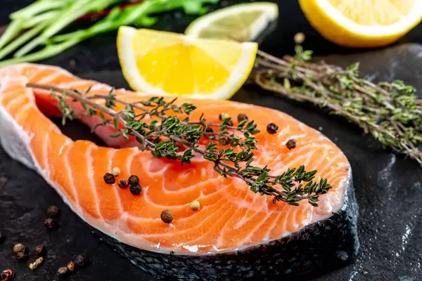 Nahaufnahme vom Lachsfleisch mit frischem Thymian und Pfefferkörner, neben Zitronenscheiben auf schwarzem Untergrund