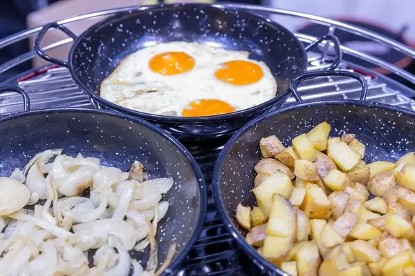 Nahaufnahme von drei Pfannen mit Bratkartoffeln, Spiegeleiern und Zwiebeln auf Metallrost