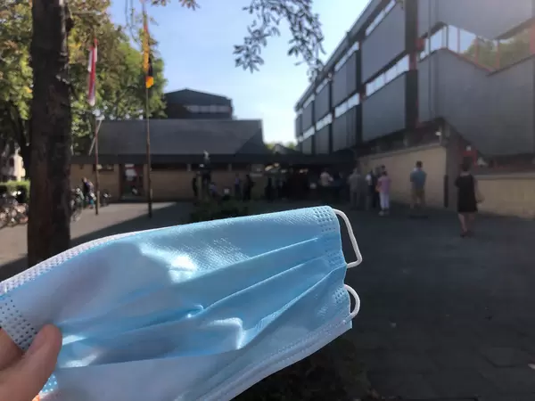 Nahaufnahme von einer OP-Maske in der Hand vor einer Schlange von Bürgern, die wählen gehen