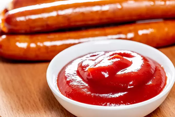 Nahaufnahme von kleiner Schale gefüllt mit Ketchup auf Holztisch mit Grillwürstchen im Hintergrund
